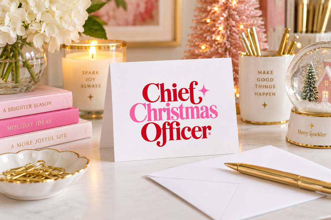 A white greeting card with the words 'Chief Christmas Officer' in pink and red lettering, styled on a tabletop with holiday decor, pink candles, a gold pen, and stationery.