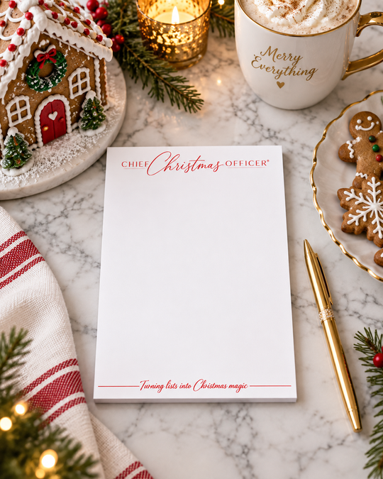 A white Christmas-themed notepad with red lettering reading 'CHIEF Christmas OFFICER' and 'Turning her into Christmas magic' is styled on a marble surface with holiday decor, including a gingerbread house, mug, candle, pen, and cookies.